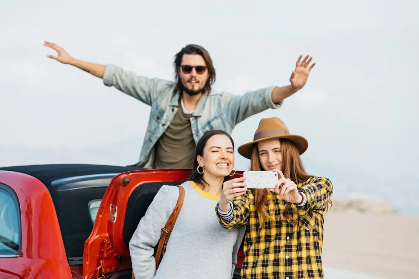 Frauen machen mit dem Smartphone ein Selfie am Kofferraum, und ein Mann lehnt sich aus einem Auto heraus.